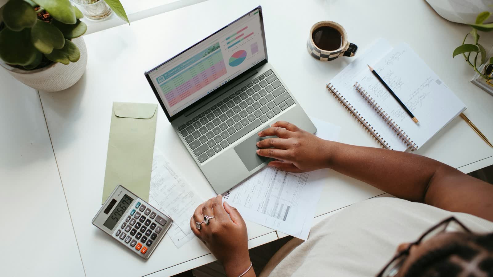A desk from above: a laptop showing a financial dashboard, a calculator, printed invoices and a notebook